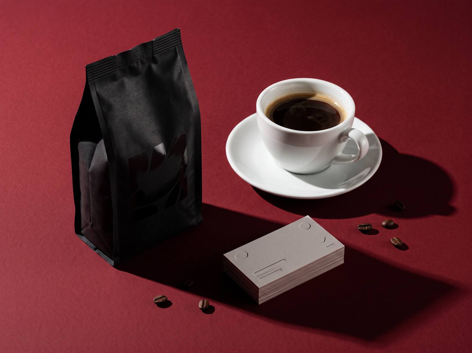 A matte black coffee bag, a cup of espresso and a stack of letterpressed business cards on a deep red table
